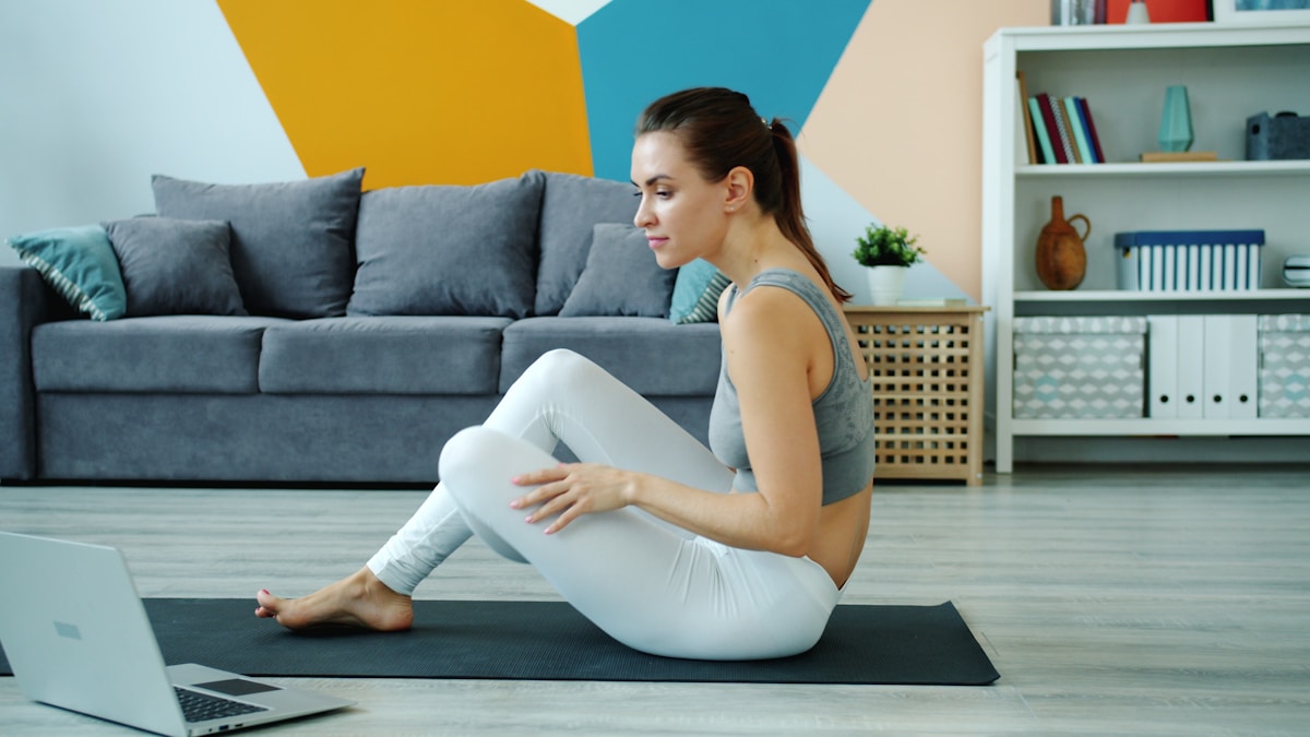 A woman on a yoga mat following a workout video on her laptop